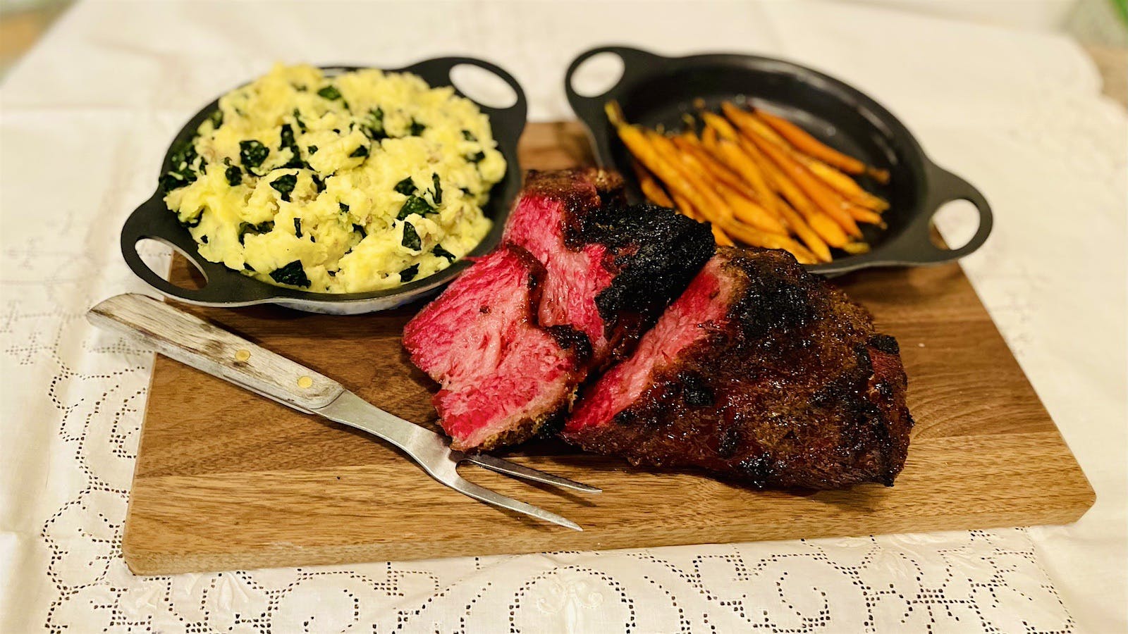 A wood cutting board with a corned beef brisket and cast iron dishes of colcannon and roasted carrots on either side